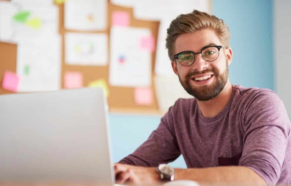A young man wearing glasses working a laptop smiles