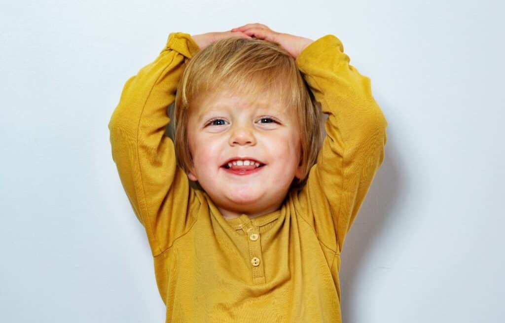 A young boy and blonde hair wearing a yellow shirt puts his hands over his head