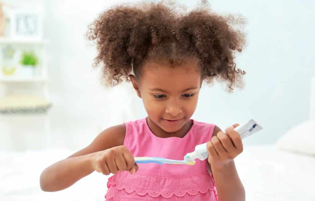 A young girl with curly hair and a pink dress squeezes toothpaste onto a toothbrush